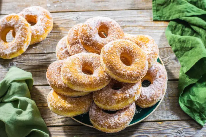 Un piatto pieno di graffe napoletane, dolci soffici e dorati, cosparse di zucchero a velo, su un tavolo di legno rustico.