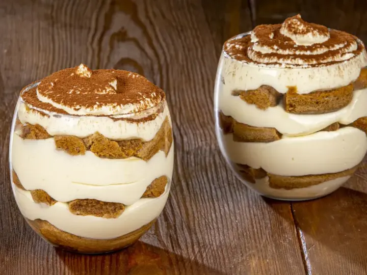 Due bicchieri di tiramisù senza uova benedetta fatto in casa, stratificato con crema e savoiardi, spolverato di cacao.