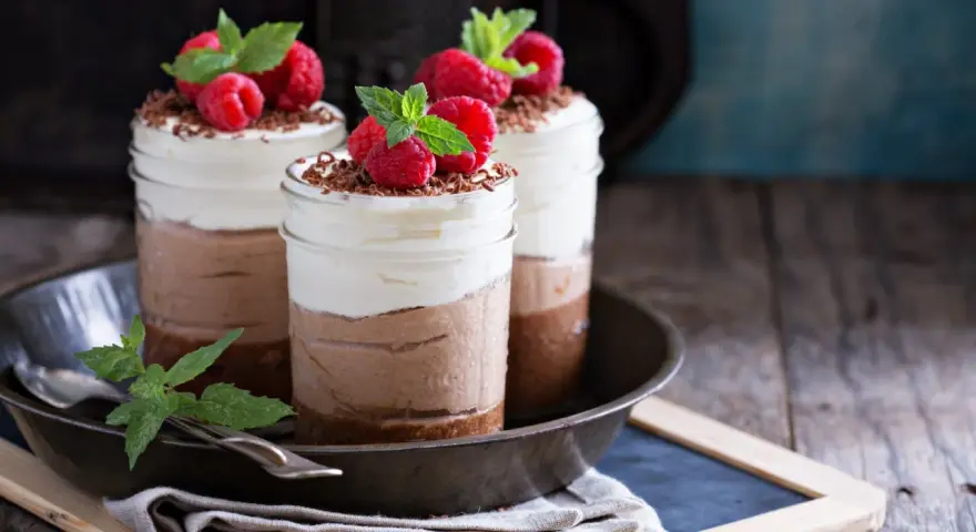 Tre deliziosi bicchierini di dolce freddo al cioccolato e panna, guarniti con lamponi freschi e scaglie di cioccolato.