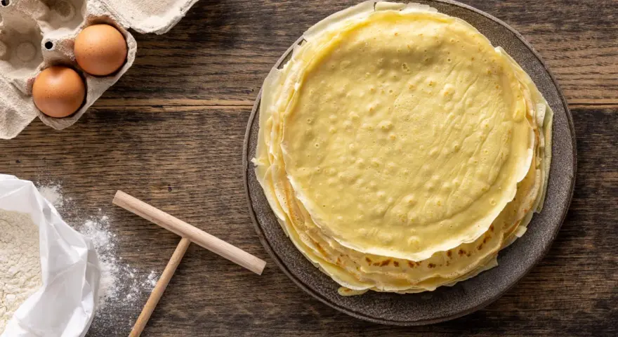 Pila di crepes dorate, pronte per essere farcite. Accanto, uova fresche e farina, ingredienti perfetti per crepes 2 uova.