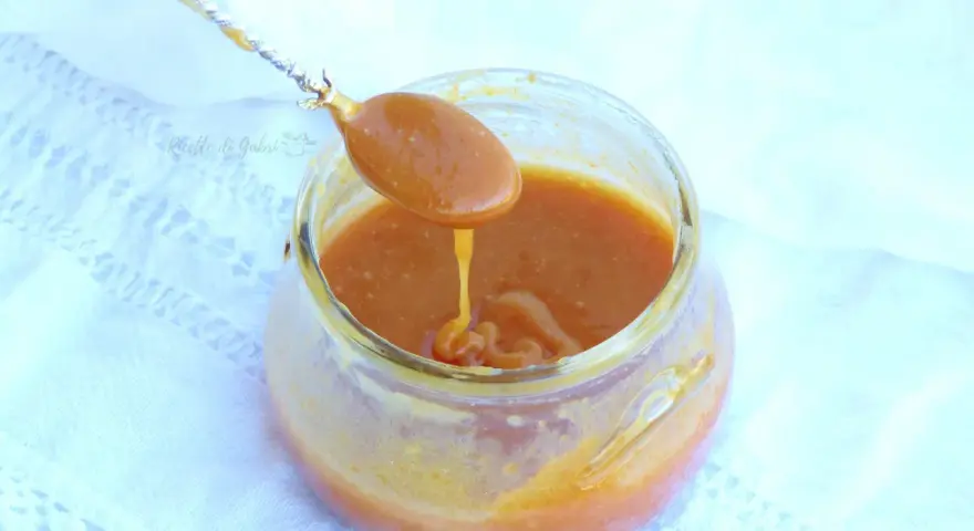 Un cucchiaio raccoglie un denso caramello salato da un barattolo di vetro. La salsa dorata cola lentamente, invitando a un assaggio goloso.