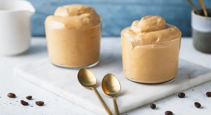Due bicchieri di soffice crema di caffè, perfetta ricetta crema di caffè senza panna e senza caffè solubile. Due cucchiaini dorati e chicchi di caffè completano la scena.