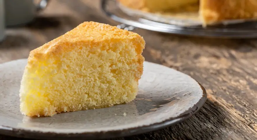 Una fetta di torta sabbiosa, soffice e dorata, su un piatto grigio. Sullo sfondo, un'altra torta intera su una gratella.