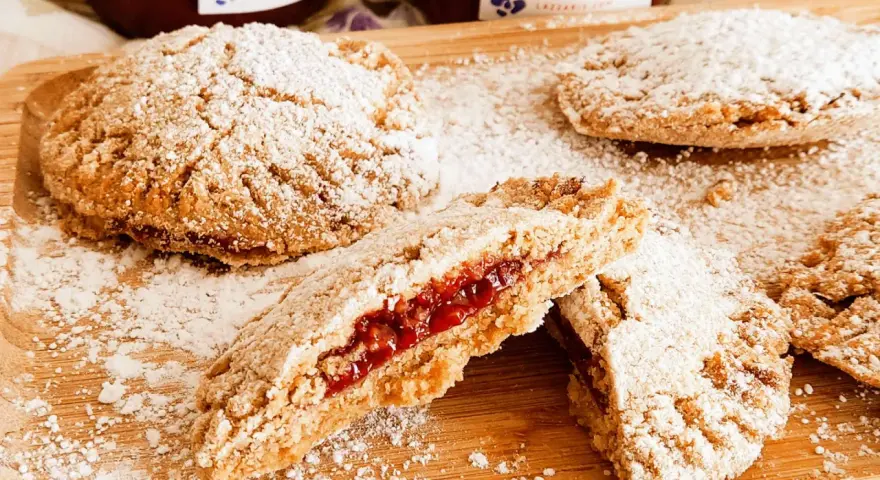 Biscotti integrali con marmellata, spolverati di zucchero a velo, su un tagliere di legno. Un morso rivela il ripieno rosso e invitante.