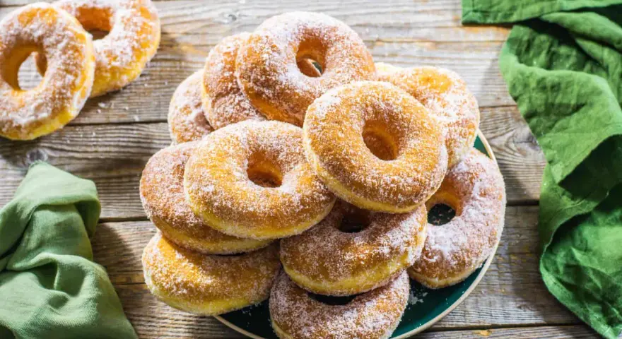 Un piatto pieno di graffe napoletane, dolci soffici e dorati, cosparse di zucchero a velo, su un tavolo di legno rustico.