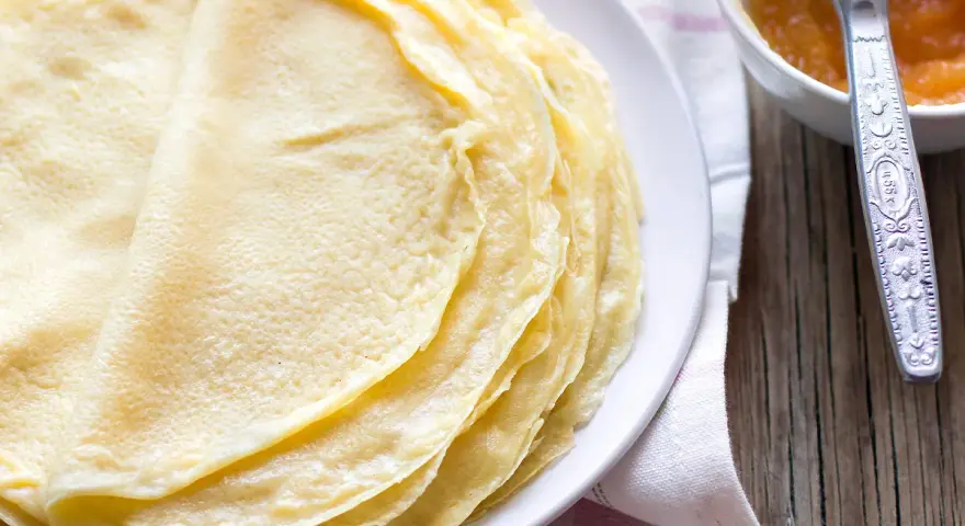 Impasto crepe dorate e sottili, pronte per essere farcite. Accanto, una ciotolina con marmellata e un coltellino.