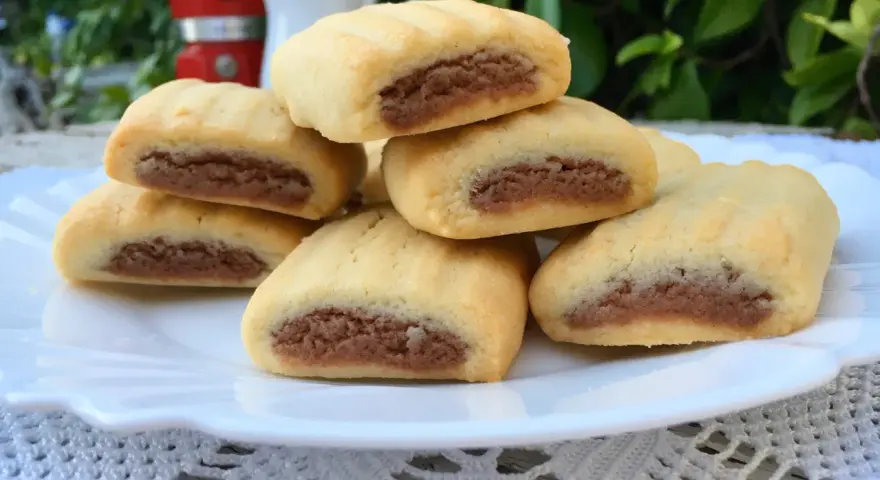 Biscotti nascondini impilati su un piatto bianco, con un ripieno scuro visibile. Sullo sfondo, una moka rossa e foglie verdi.