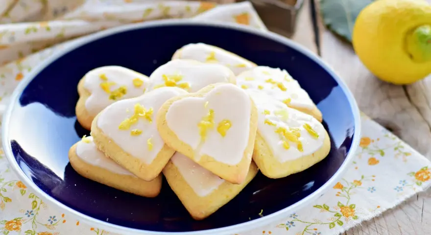Cuore di biscotti al limone glassati, decorati con scorza di limone, su un piatto blu.