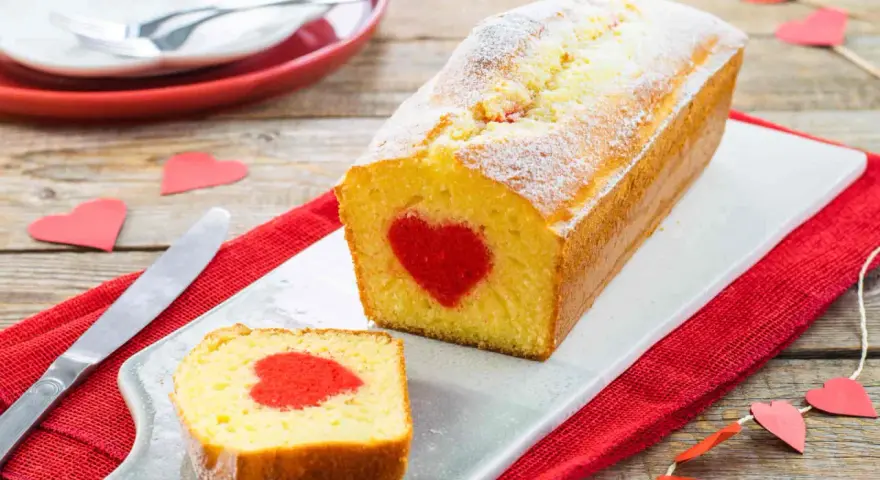 Plumcake con cuore rosso al centro, cosparso di zucchero a velo. Una fetta tagliata rivela la sorpresa. Decorazioni a forma di cuore completano l'atmosfera romantica.