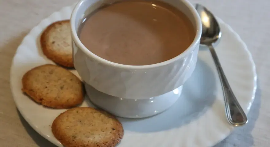 Tazza di crema di cogne fumante con biscotti secchi su un piattino bianco, accompagnata da un cucchiaino.
