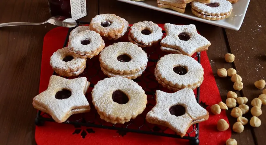 Deliziosi biscotti Linzer, a forma di stella e tondi, ripieni di marmellata e spolverati di zucchero a velo, su una griglia nera.