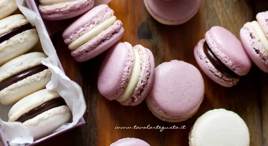 Deliziosi macaron dolci rosa e bianchi, alcuni con ripieno al cioccolato, sparsi su un tagliere di legno.