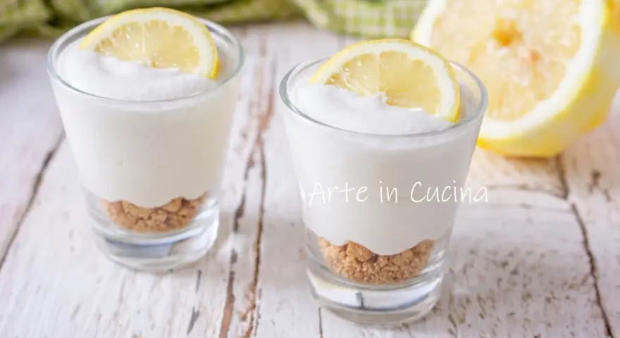 Due bicchierini di mousse al limone con base croccante e fetta di limone, su un tavolo di legno.