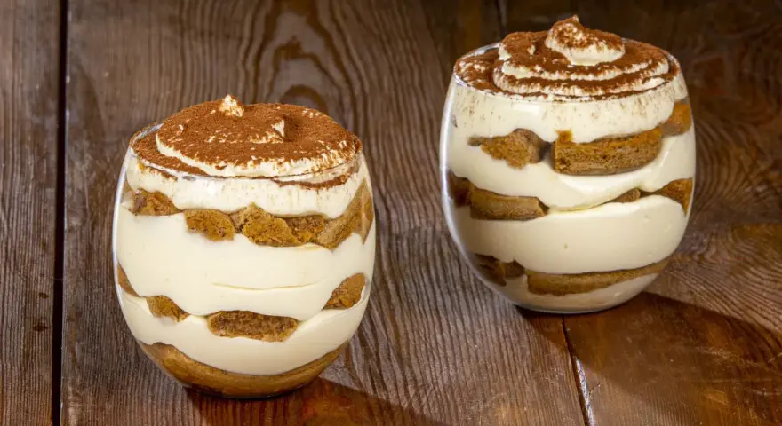 Due bicchieri di tiramisù senza uova benedetta fatto in casa, stratificato con crema e savoiardi, spolverato di cacao.