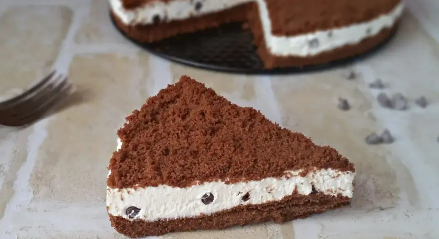 Una fetta di torta al cioccolato con ripieno cremoso al mascarpone e biscotti sbriciolati, decorata con gocce di cioccolato.