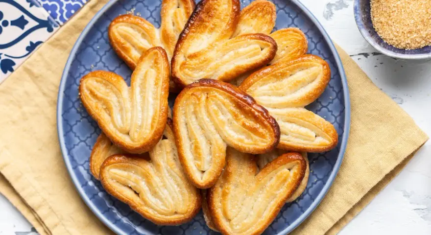 Un piatto blu pieno di croccanti ventagli di pasta sfoglia, dorati e zuccherati, pronti per essere gustati.