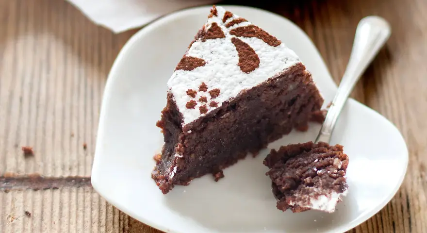 Una fetta di torta caprese ricetta facile, cosparsa di zucchero a velo e cacao, su un piatto bianco a forma di cuore.