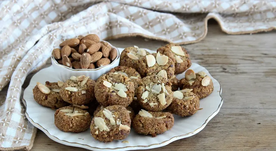 Biscotti senza zucchero, dorati e croccanti, guarniti con mandorle a lamelle. Accanto, una ciotola piena di mandorle intere.