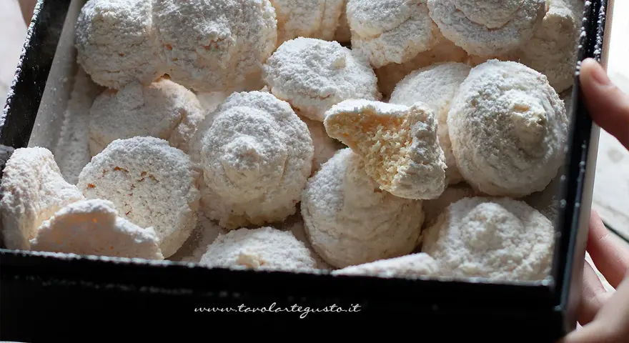 Scatola piena di soffici dolcetti al cocco, spolverati di zucchero a velo. Uno è spezzato, mostrando l'interno friabile.
