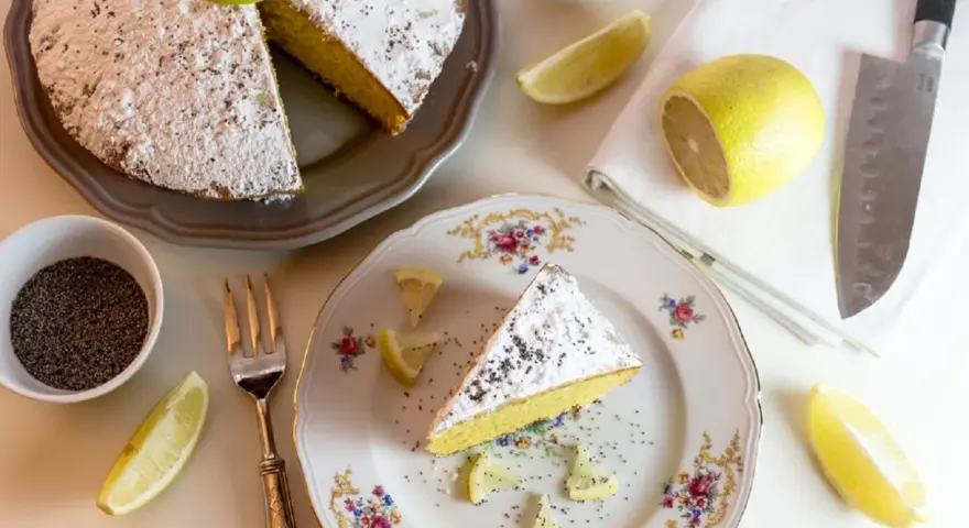 Una fetta di torta al limone cosparsa di zucchero a velo e semi di papavero, guarnita con spicchi di limone fresco.