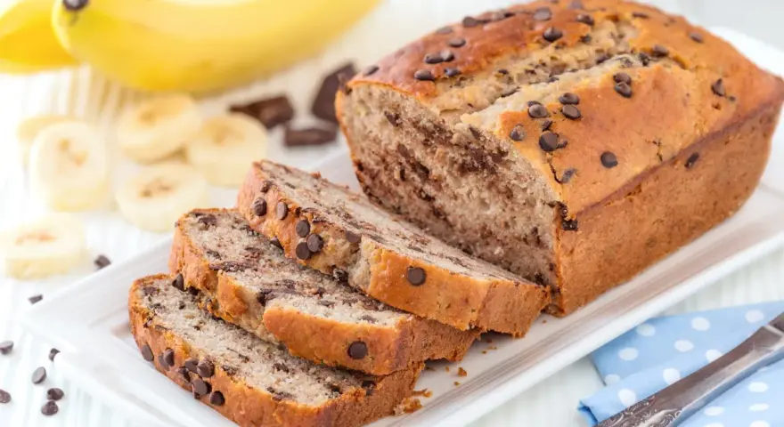 Fette di plumcake banana e cioccolato su un piatto bianco, con banane e gocce di cioccolato sullo sfondo.