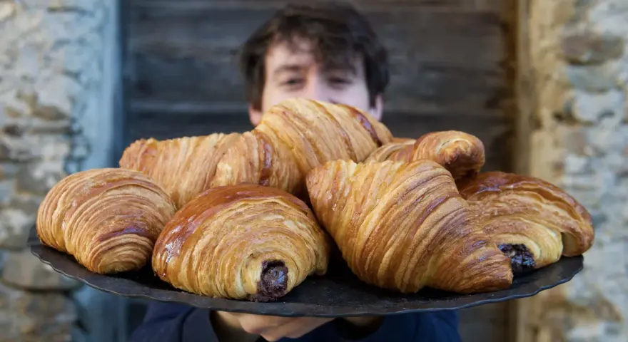 Un ragazzo sorride dietro un vassoio pieno di croissant brioche dorati e fragranti, alcuni con ripieno di cioccolato.