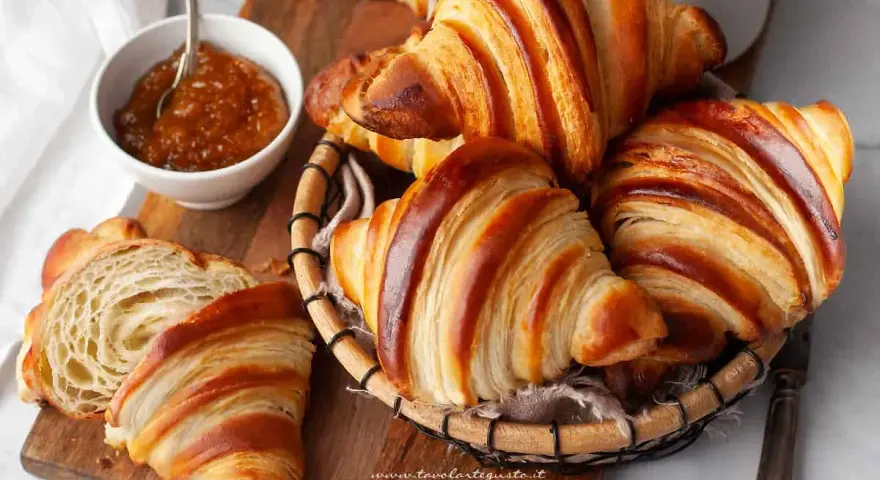 Croissant dorati e sfogliati, un invito a scoprire la loro origine. Marmellata in ciotola e una tazza bianca completano la scena.