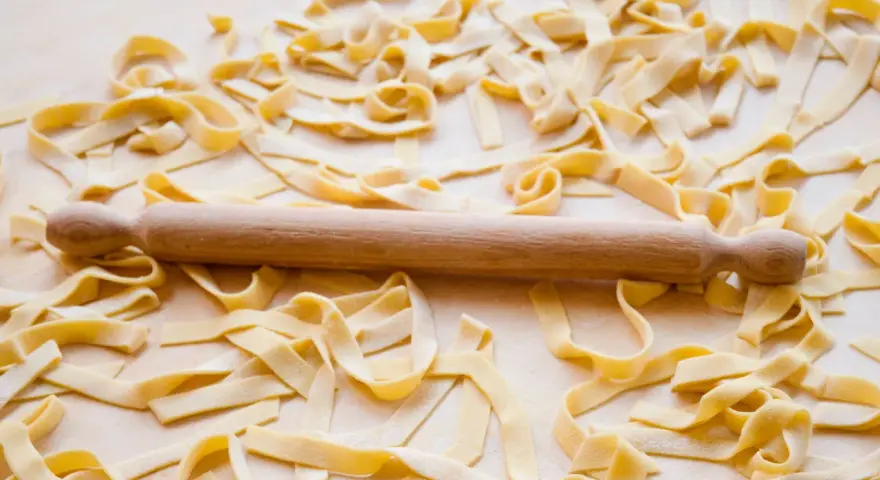 Fettuccine fresche sparse su un piano di lavoro, con un mattarello in legno. La farina, che è all-purpose flour, è visibile sulla pasta.