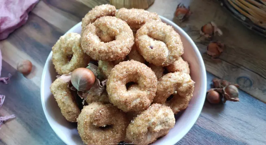 Ciambelline al vino cosparse di zucchero, con nocciole sparse sul tavolo di legno.