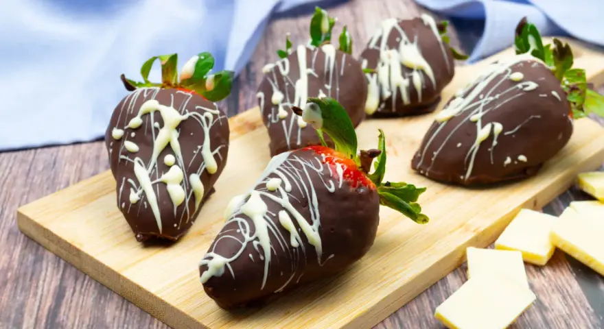 Fragole ricoperte di cioccolato fondente e decorate con cioccolato bianco. Un delizioso assaggio di frutta al cioccolato su un tagliere di legno.