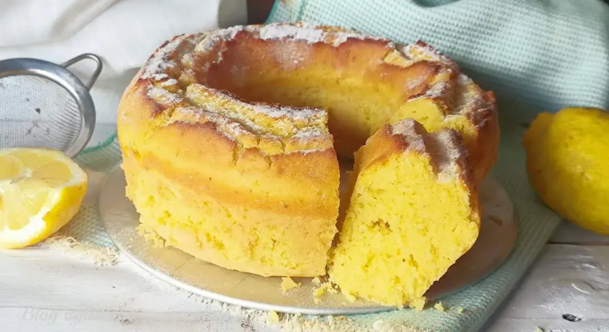 Torta soffice e dorata, spolverata di zucchero a velo, preparata con dolci con eritritolo. Accanto, limoni freschi e un setaccio.