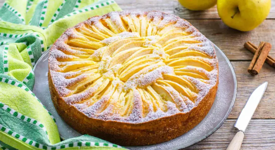 Torta di mele con yogurt, decorata con fettine di mela e zucchero a velo, su un piatto grigio, accanto a un tovagliolo a righe verdi e bianche.