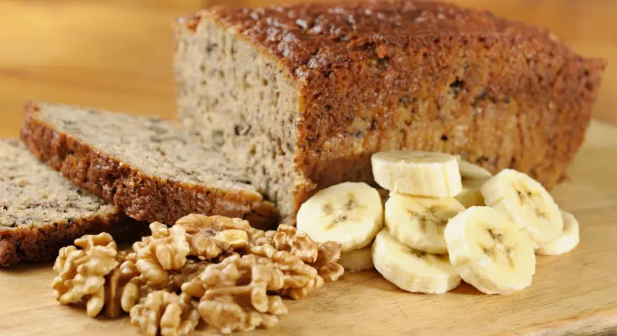 Fetta di banana bread senza uova, con noci e banane a fette, su tagliere di legno.