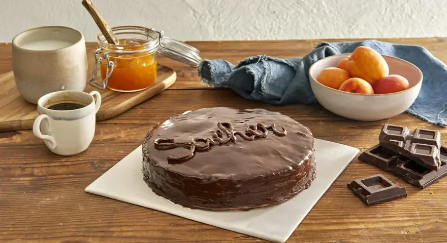 Torta Sacher, un classico viennese, con la sua glassa lucida e il nome scritto in cioccolato. Accanto, albicocche, marmellata e caffè, ingredienti che evocano le sue origini.