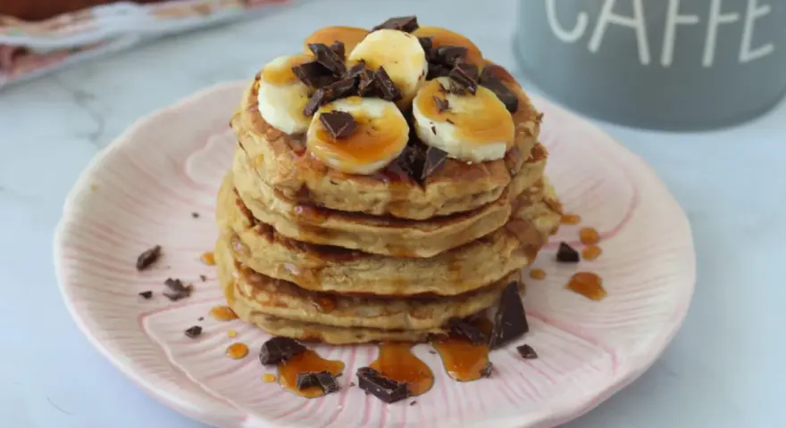 Pancake light con banana, cioccolato e sciroppo d'acero su un piatto rosa.