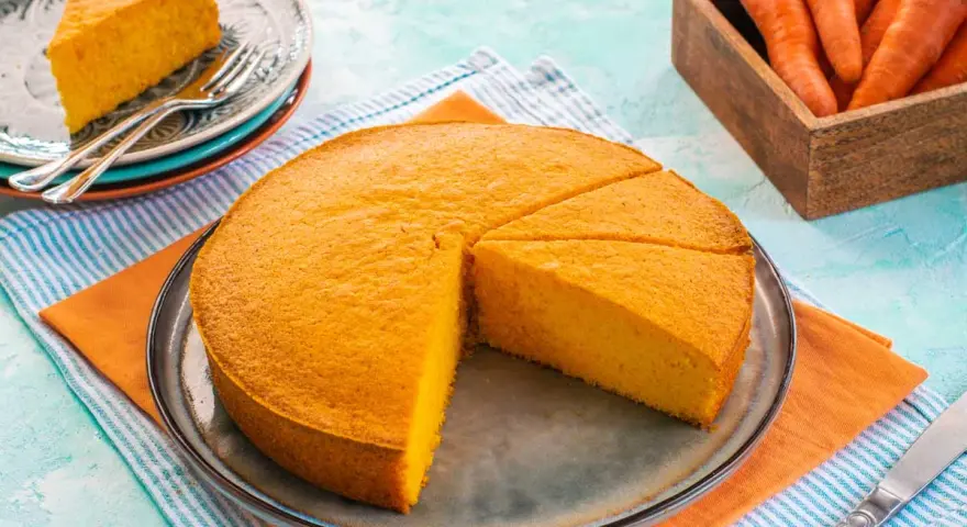 Una deliziosa torta di carote, appena sfornata, è tagliata a fette su un piatto. Accanto, un cesto di carote fresche.