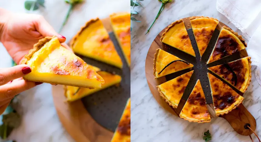 Una mano prende una fetta di flan parisien dorato. La torta intera, tagliata a spicchi, è su un tagliere di legno, decorata con un fiore e un rametto di eucalipto.