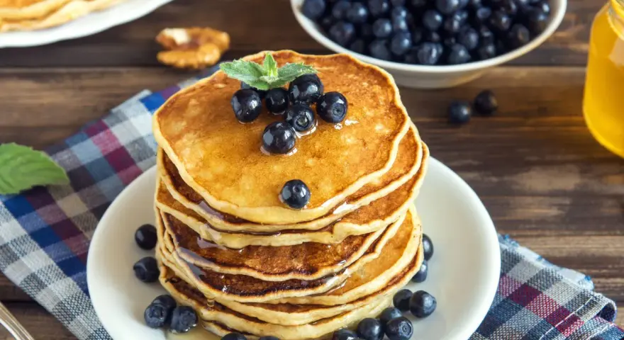 Pila di pancake vegani dorati, guarniti con mirtilli freschi e sciroppo d'acero, su un piatto bianco, con una ciotola di mirtilli e una bottiglia di sciroppo sullo sfondo.