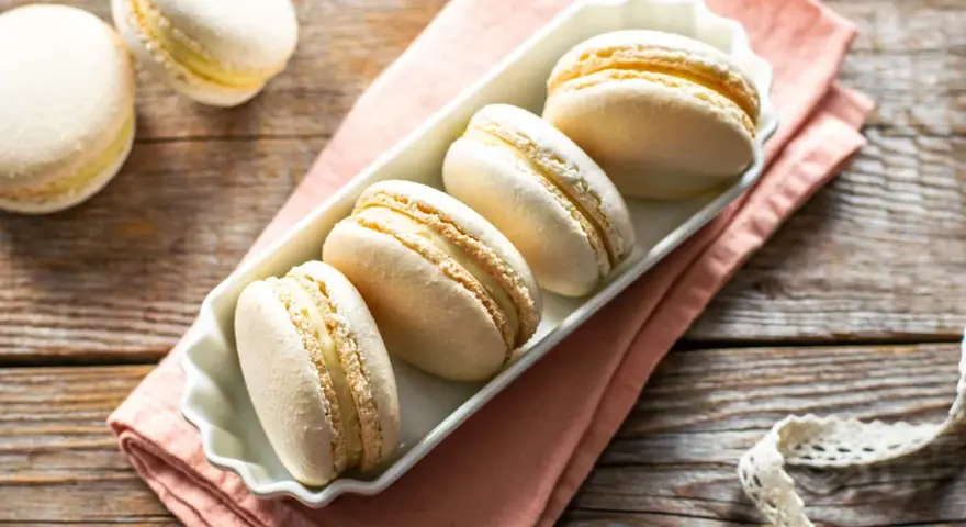 Macarons bianchi e dorati, perfetti esempi di come sono fatti i macarons, con ripieno cremoso, su un piatto bianco e tovagliolo rosa.