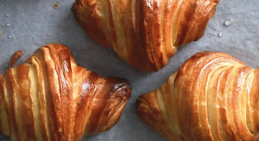 Tre cornetti dorati e sfogliati, appena sfornati, con gocce di condensa sulla carta da forno.