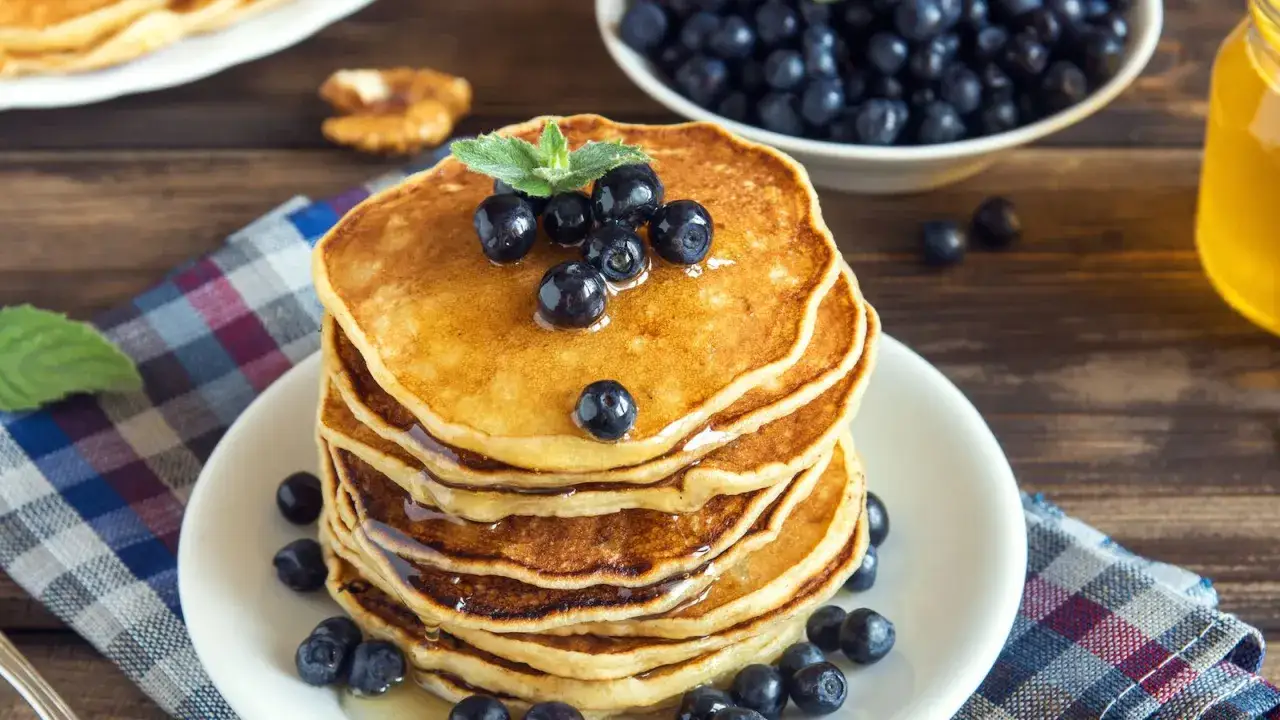 Pila di pancake vegani dorati, guarniti con mirtilli freschi e sciroppo d'acero. Sullo sfondo, una ciotola di mirtilli e un barattolo di miele.