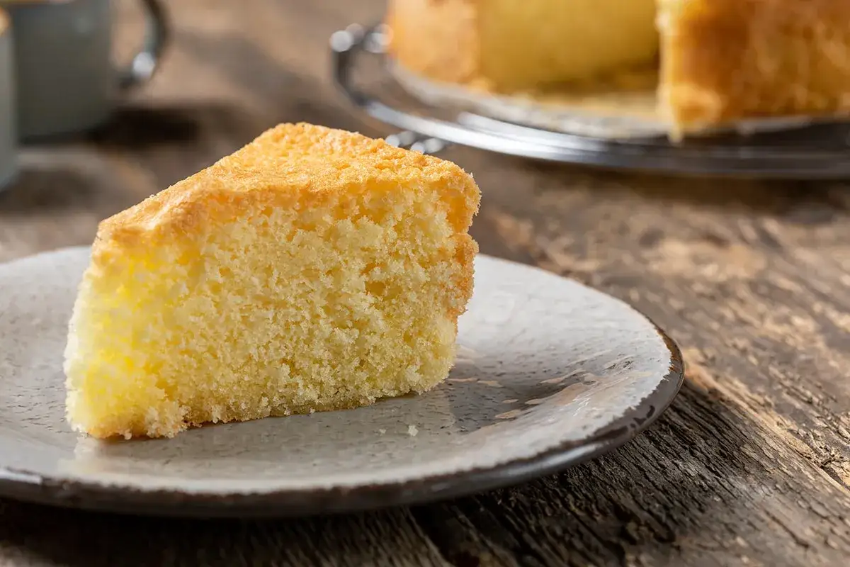 Una fetta di torta sabbiosa, soffice e dorata, su un piatto grigio. Sullo sfondo, un'altra torta intera su una gratella.