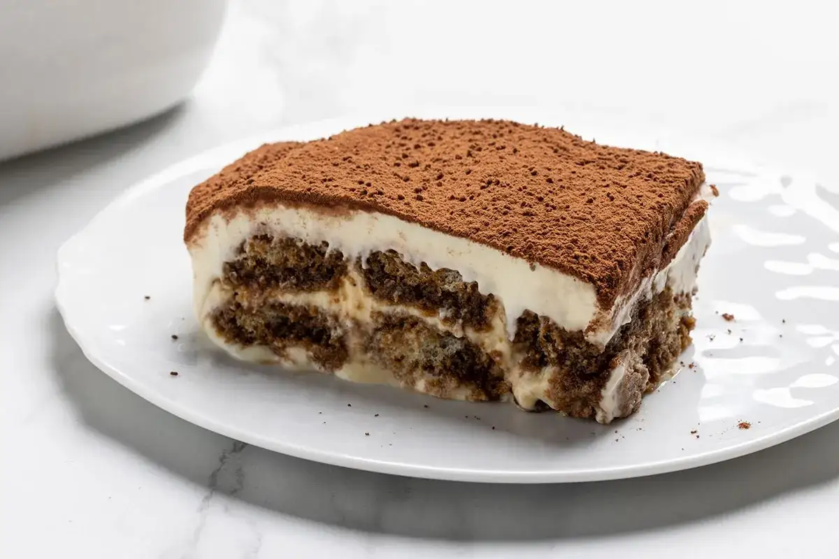 Una fetta di tiramisù su un piatto bianco, con strati di savoiardi inzuppati e crema al mascarpone, spolverata di cacao. La ricetta tiramisù 500 g mascarpone è un classico.