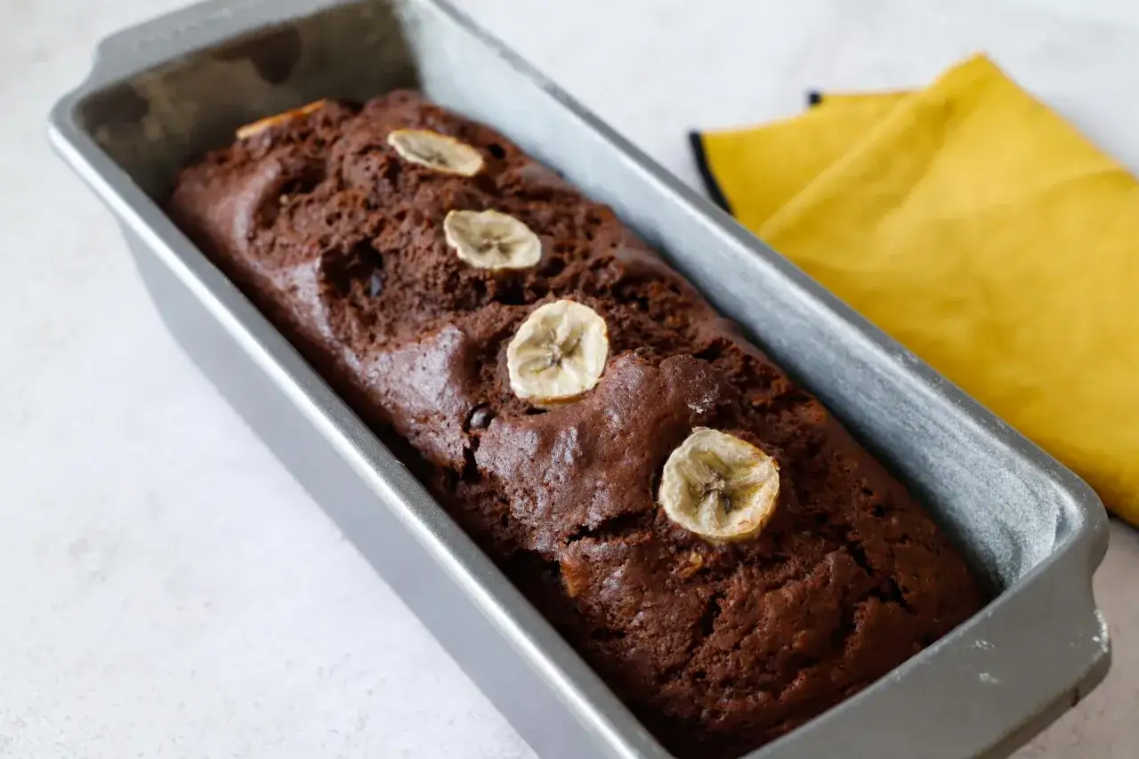 Plumcake banana e cioccolato appena sfornato, decorato con fettine di banana, in una teglia metallica.