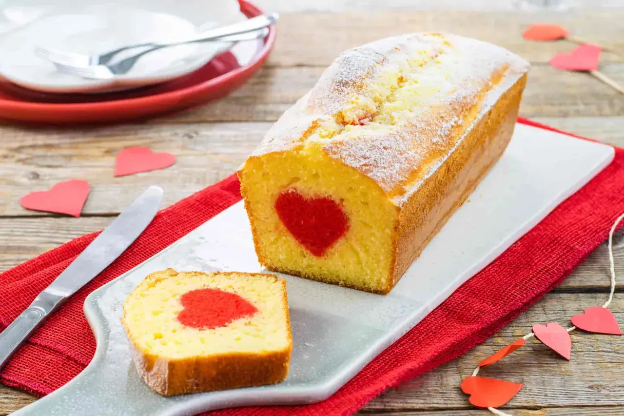 Plumcake con cuore rosso al centro, una fetta tagliata mostra la sorpresa. Decorazioni a forma di cuore e un coltello completano la scena.