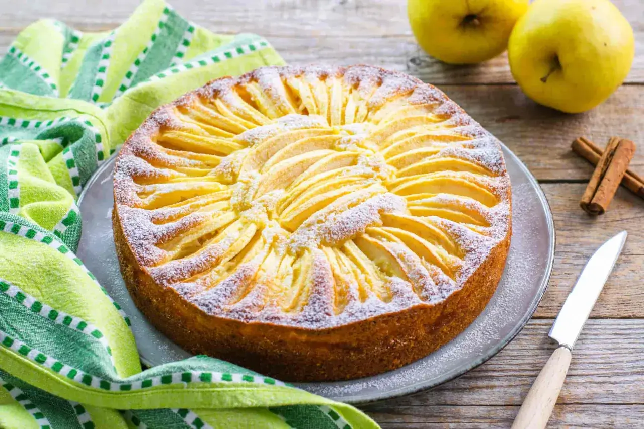 Torta di mele con yogurt, decorata con fettine di mela e zucchero a velo, su un piatto grigio, accanto a un tovagliolo verde e cannella.