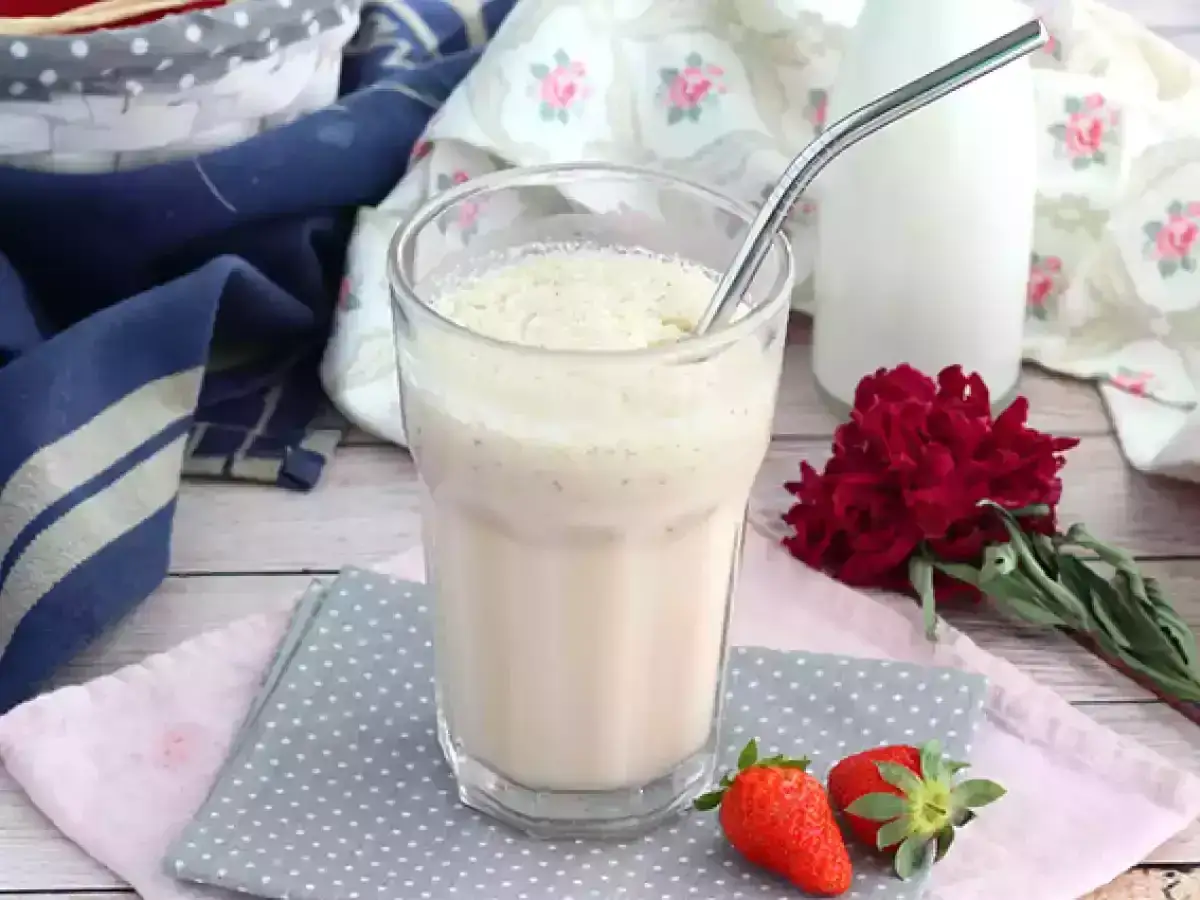 Delizioso milkshake alla vaniglia, perfetto per una milkshake ricetta veloce. Due fragole fresche e un fiore rosso completano la scena.