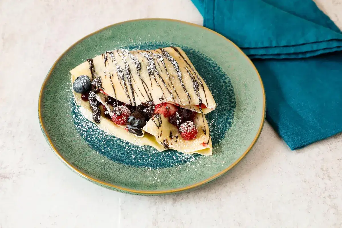 Crepes 2 uova con frutti di bosco e cioccolato, spolverate di zucchero a velo su un piatto verde.