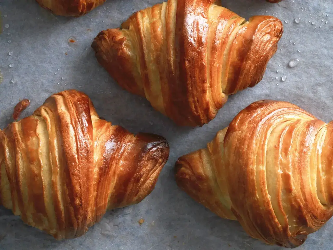 Tre croissant brioche dorati e sfogliati, appena sfornati, su carta da forno con gocce d'acqua.