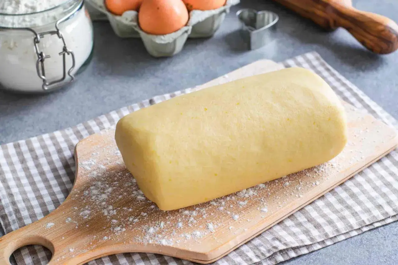 Rotolo di pasta frolla con strutto, pronto per essere trasformato in deliziosi biscotti. Ingredienti freschi e un utensile a forma di cuore sullo sfondo.